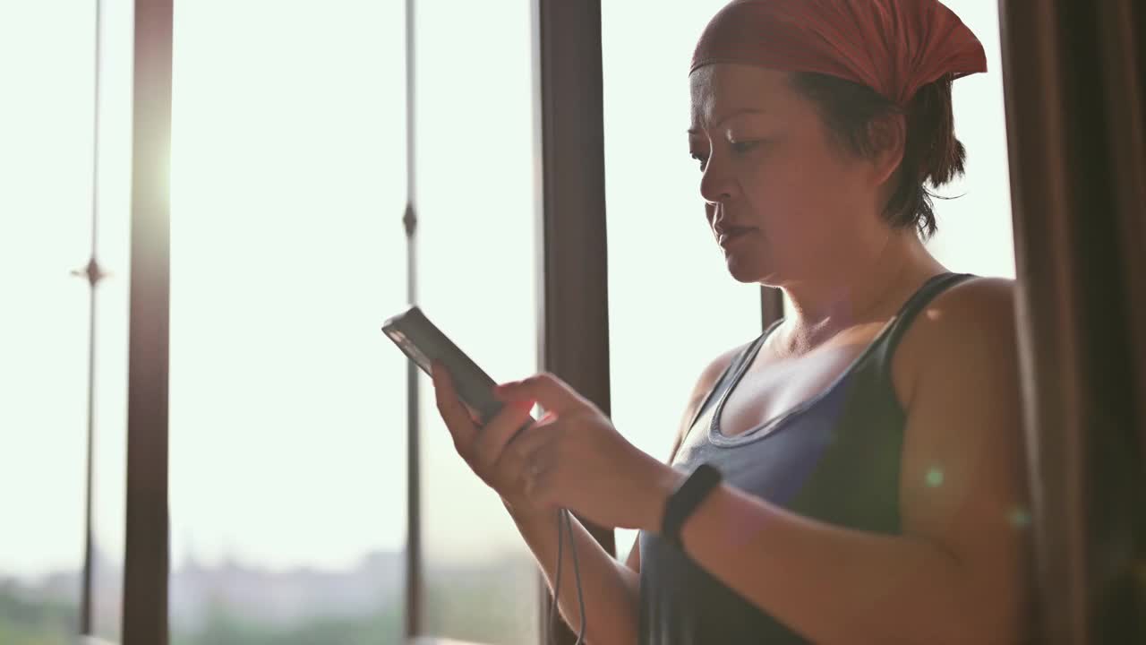 亚洲华人中年女性戴着头巾，早上在阳光明媚的窗户旁用智能手机查看健身追踪器视频下载