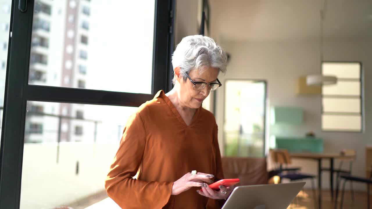 高级女性在家使用笔记本电脑和智能手机视频下载