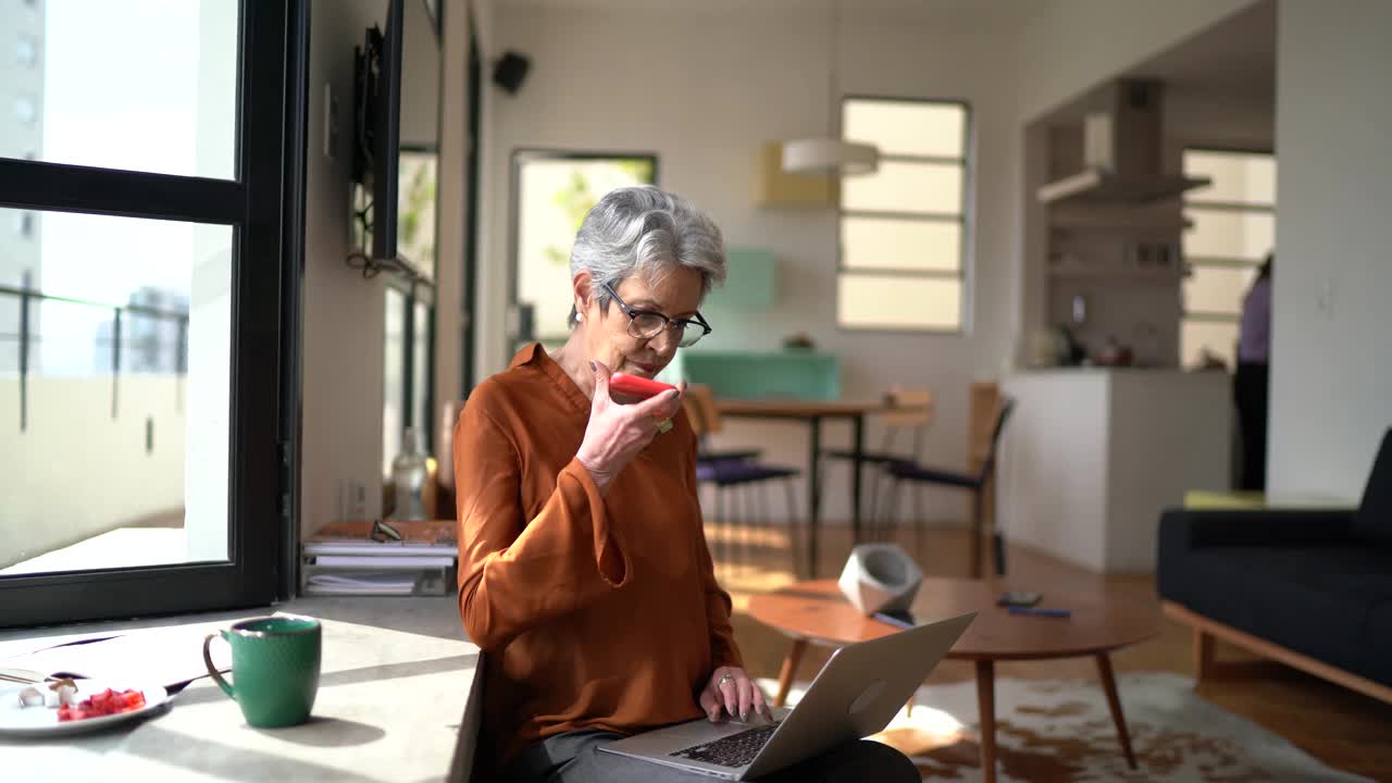 高级女性在家工作，使用笔记本电脑，用智能手机发送音频信息视频下载