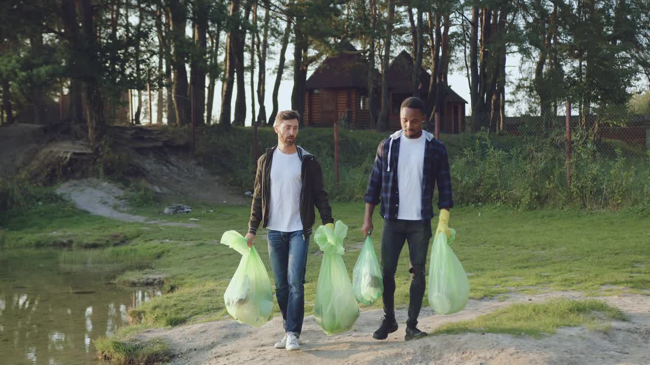 英俊满意的混血儿年轻男子背着塑料袋在湖边附近清理了周围的垃圾，正面的观点视频下载