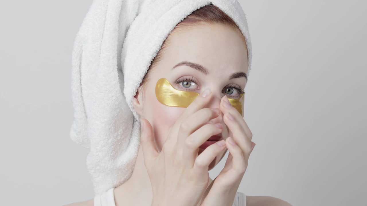 美丽微笑的女人头上的毛巾，在家庭水疗过程中应用金色的眼罩视频素材