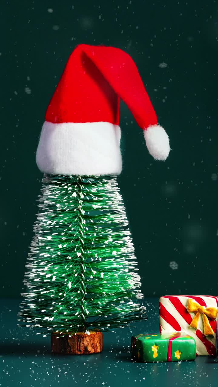圣诞快乐和新年快乐雪花飘落，绿色松树上的圣诞帽和深绿色背景上的礼盒。节日庆祝贺卡视频下载