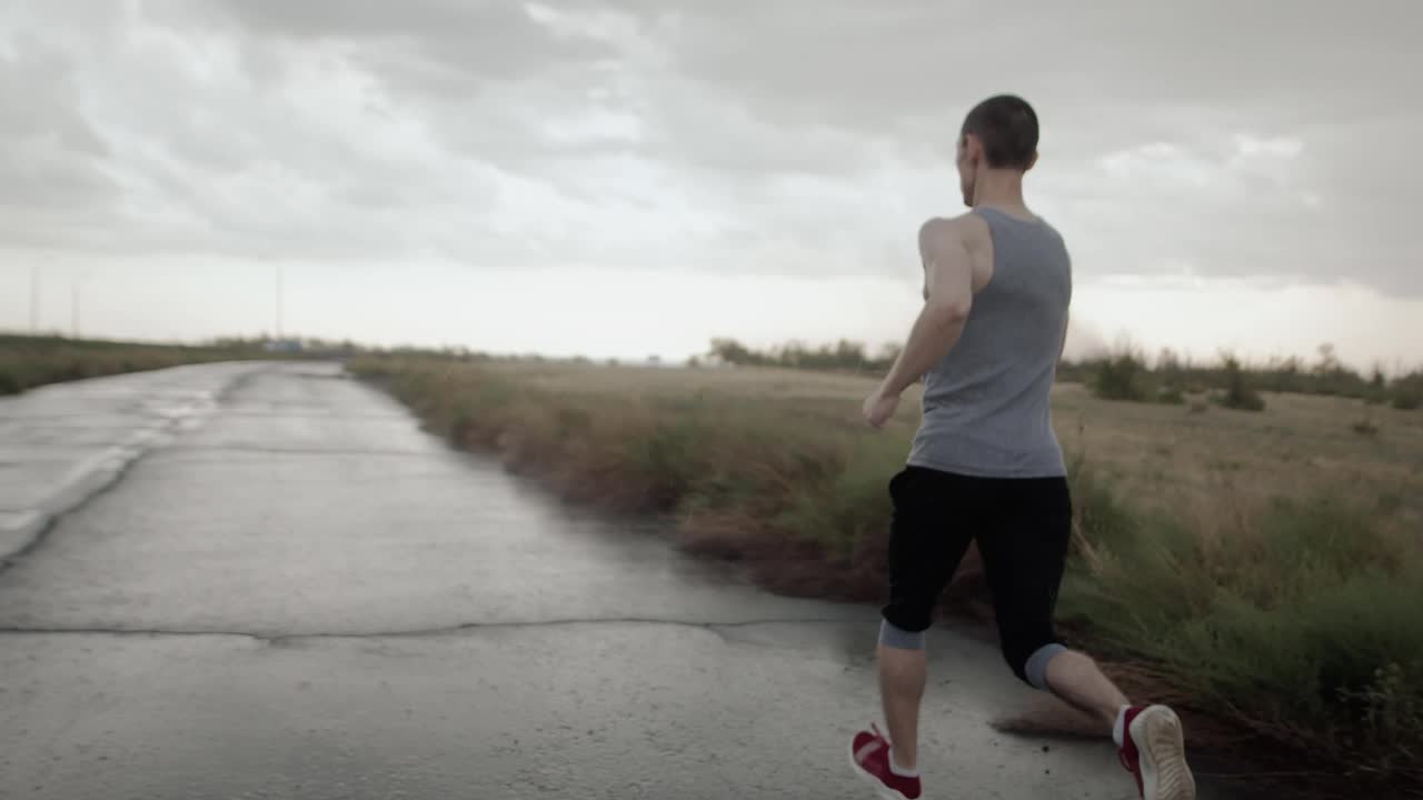 运动员身材的男子在田野和阴天的背景下沿着沥青公路奔跑，向后看。动机视频素材