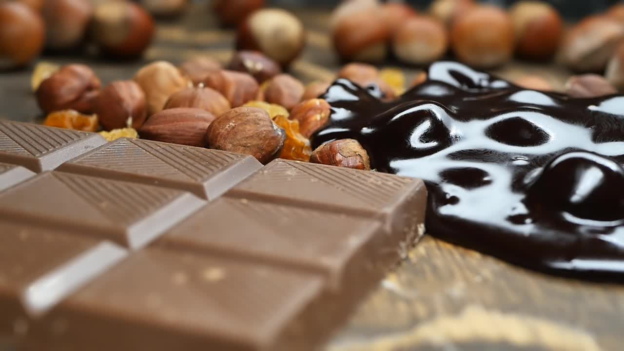 将黑苦巧克力倒在整个榛子和葡萄干上，放在巧克力棒旁边。多莉，慢镜头。木制背景。视频素材