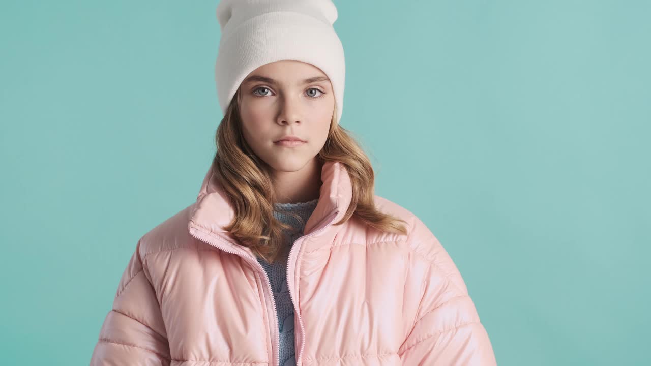 漂亮凉爽的少女穿着粉红色的羽绒服和帽子在蓝色背景下看起来严重孤立。时尚的外观视频下载