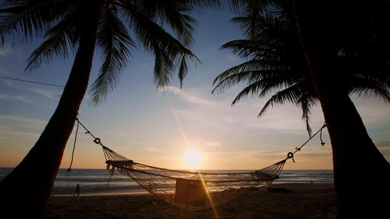 泰国普吉岛的海滩。日落时分，热带海滩上吊床和棕榈树的剪影。吊床上的棕榈树和日落眩目附近的海洋，海洋，天空，海岸，沙滩。视频素材
