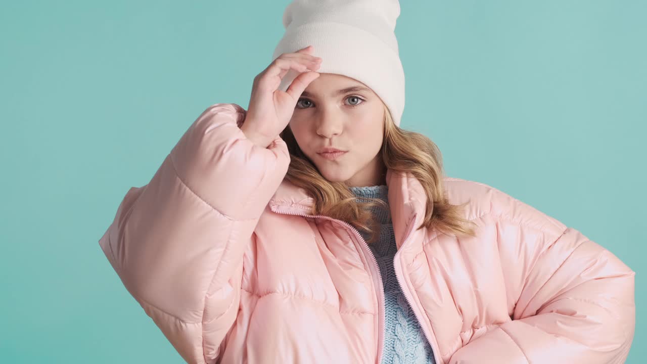 非常有趣的少女穿着粉红色的羽绒服，戴着帽子遮住脸，在蓝色的背景下看着周围的镜头视频下载