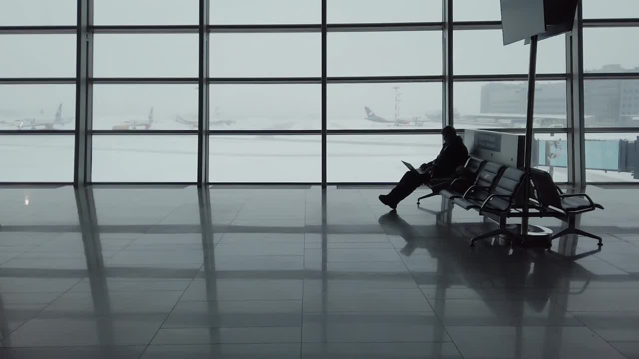 在冬天的雪天里，一名男子坐在机场候机室里，用笔记本电脑和手机拍照，背景是飞机视频素材
