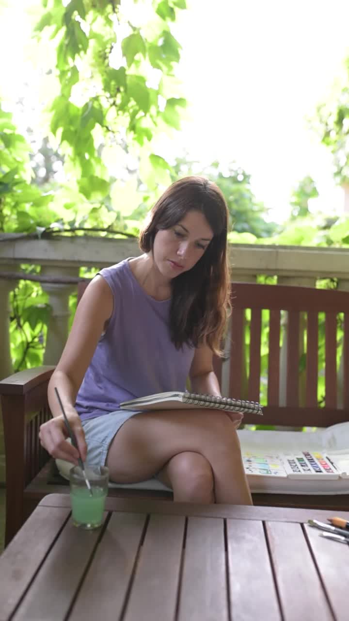 一个年轻的白人黑发女人坐在花园长椅上，她的腿伸在长凳上，在她的笔记本上画水彩画。视频下载