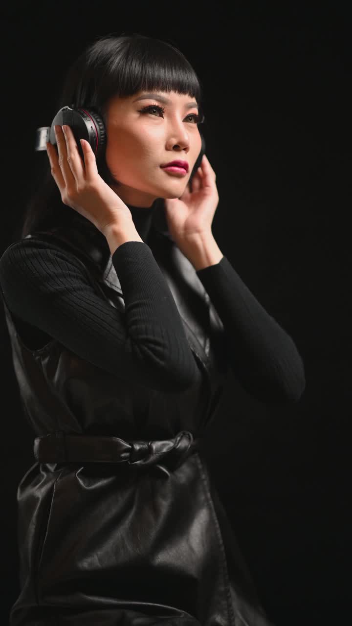 亚洲的中国女时装模特穿着皮夹克听着耳机在摄影棚肖像眼睛关闭黑色背景视频素材