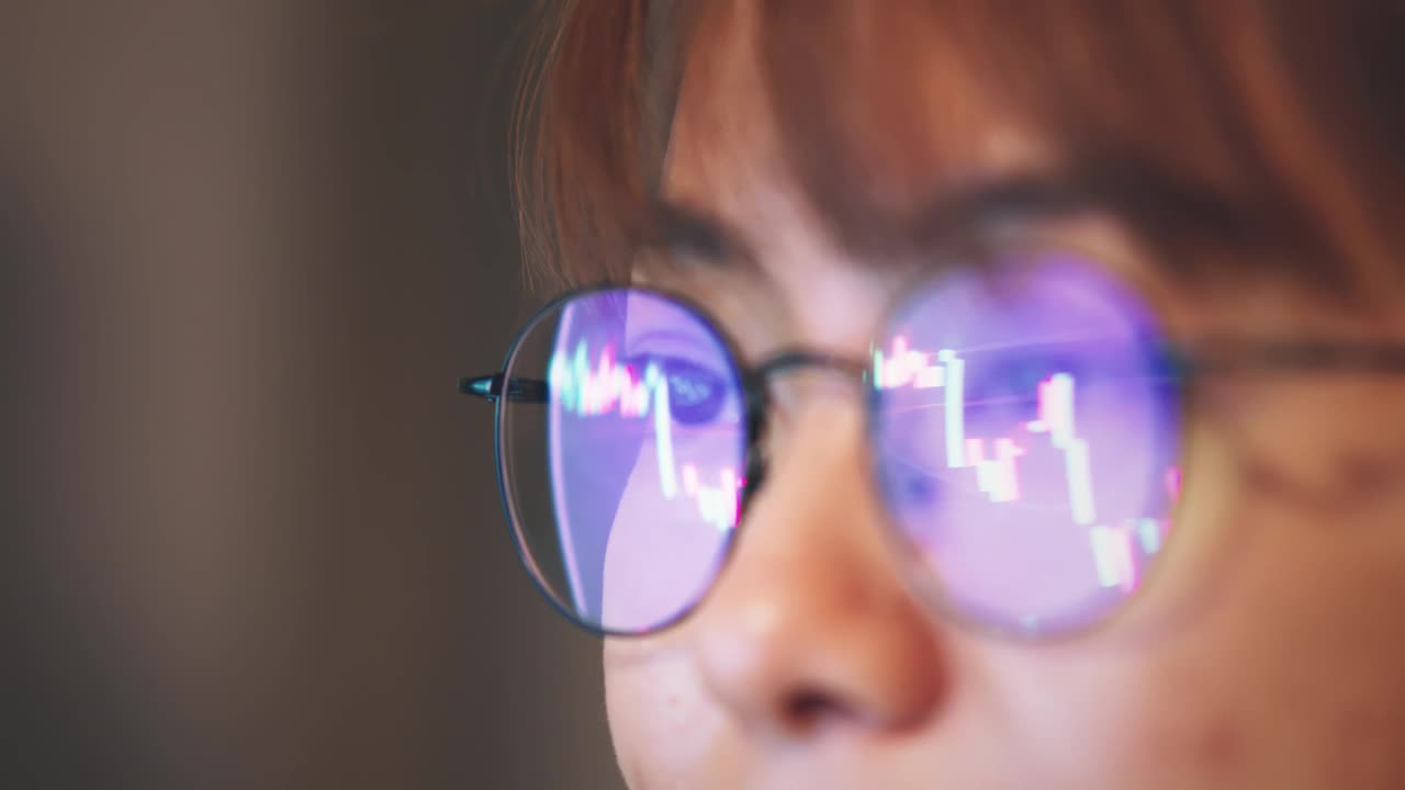 戴着眼镜的女性眼睛深夜在笔记本电脑前滚动。作为一名程序员，她在智能手机上查看比特币的数字交易价格图表。视频素材