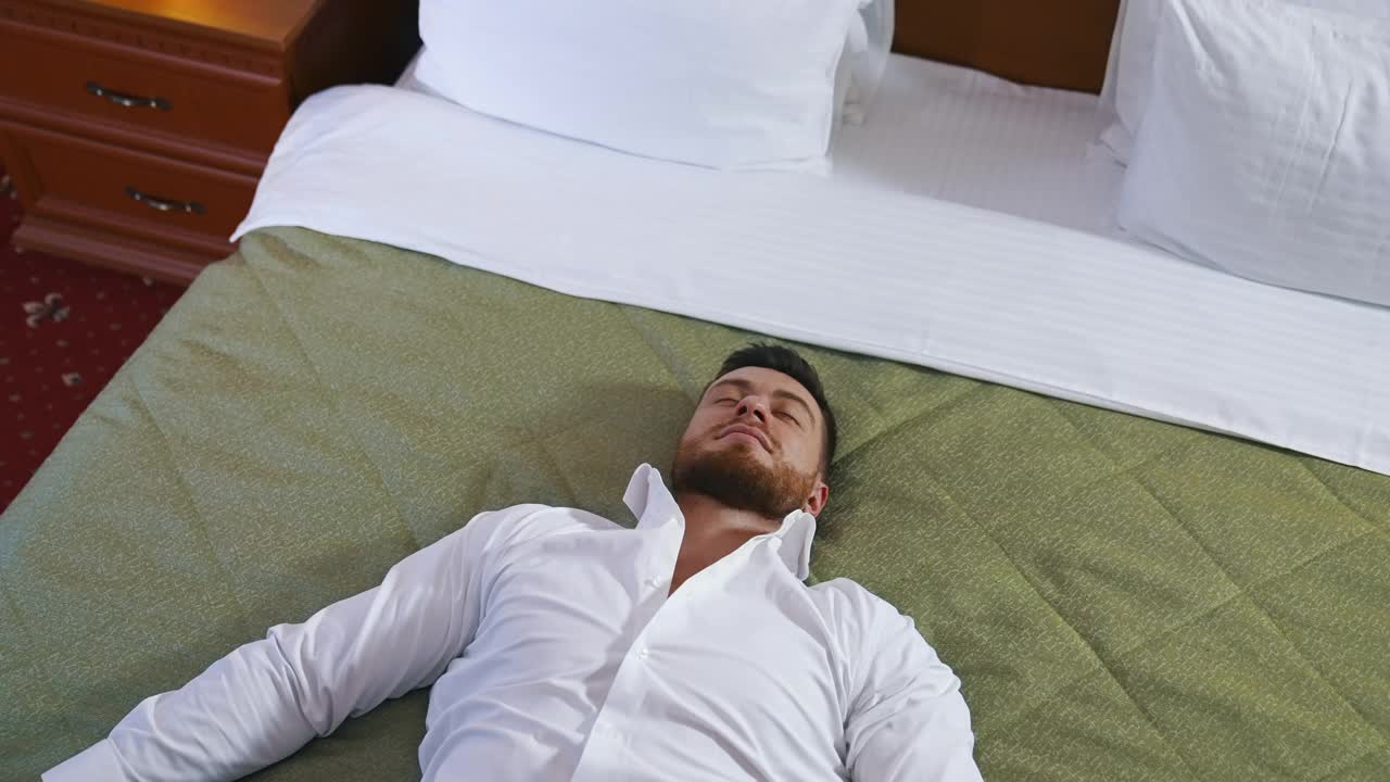 年轻的商人躺在床上休息。一个穿着白衬衫的英俊男子独自躺在酒店房间的床上休息。出差期间休假。视频下载