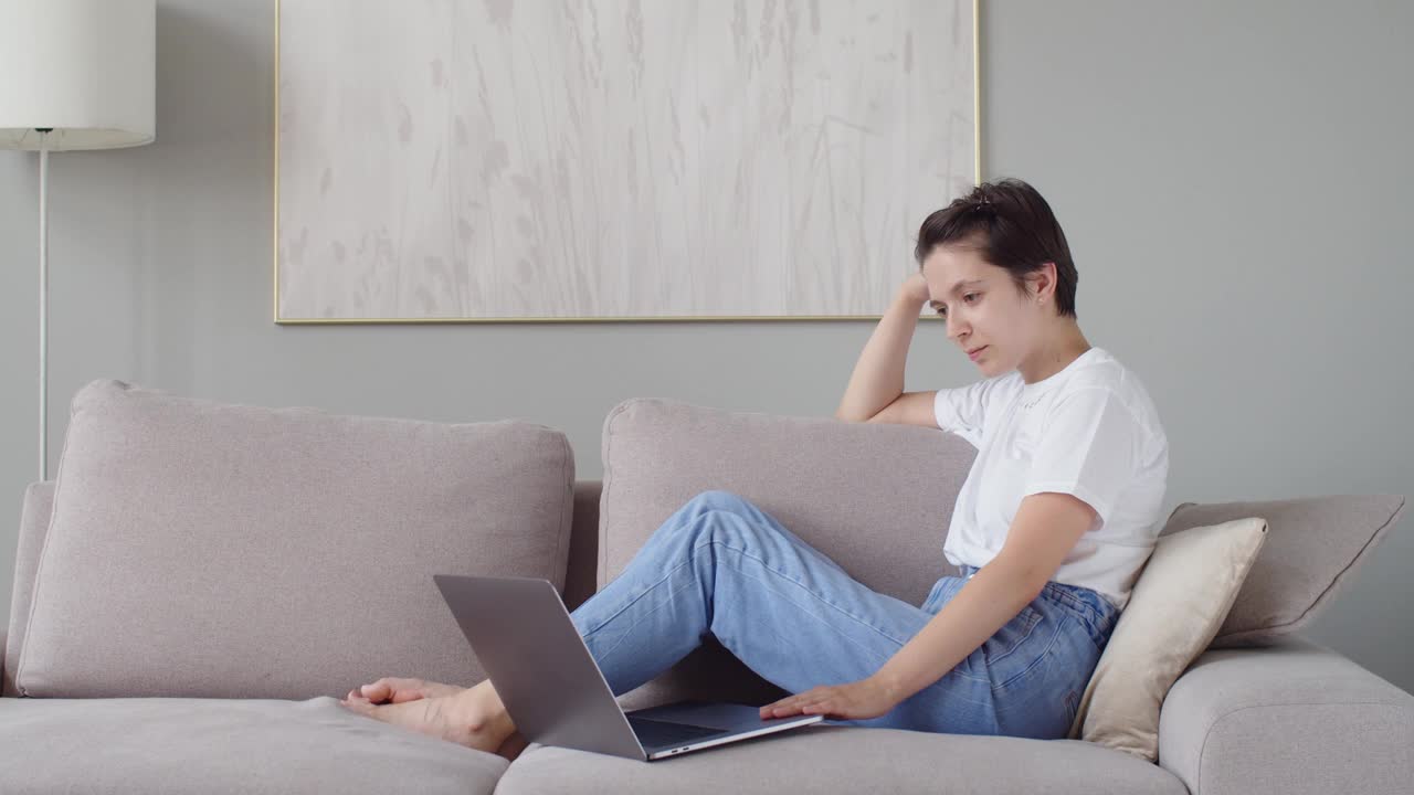 年轻女士坐在沙发上用笔记本电脑用快乐的表情复制空间文本。微笑的女人用慢动作和朋友聊天打发闲暇时间。技术交流视频下载