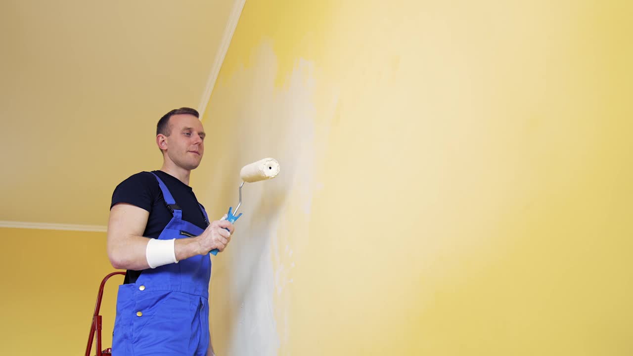 房间改造。画墙的年轻人的肖像。工人从室内的梯子上用油漆滚筒给墙壁刷新颜色。视频素材