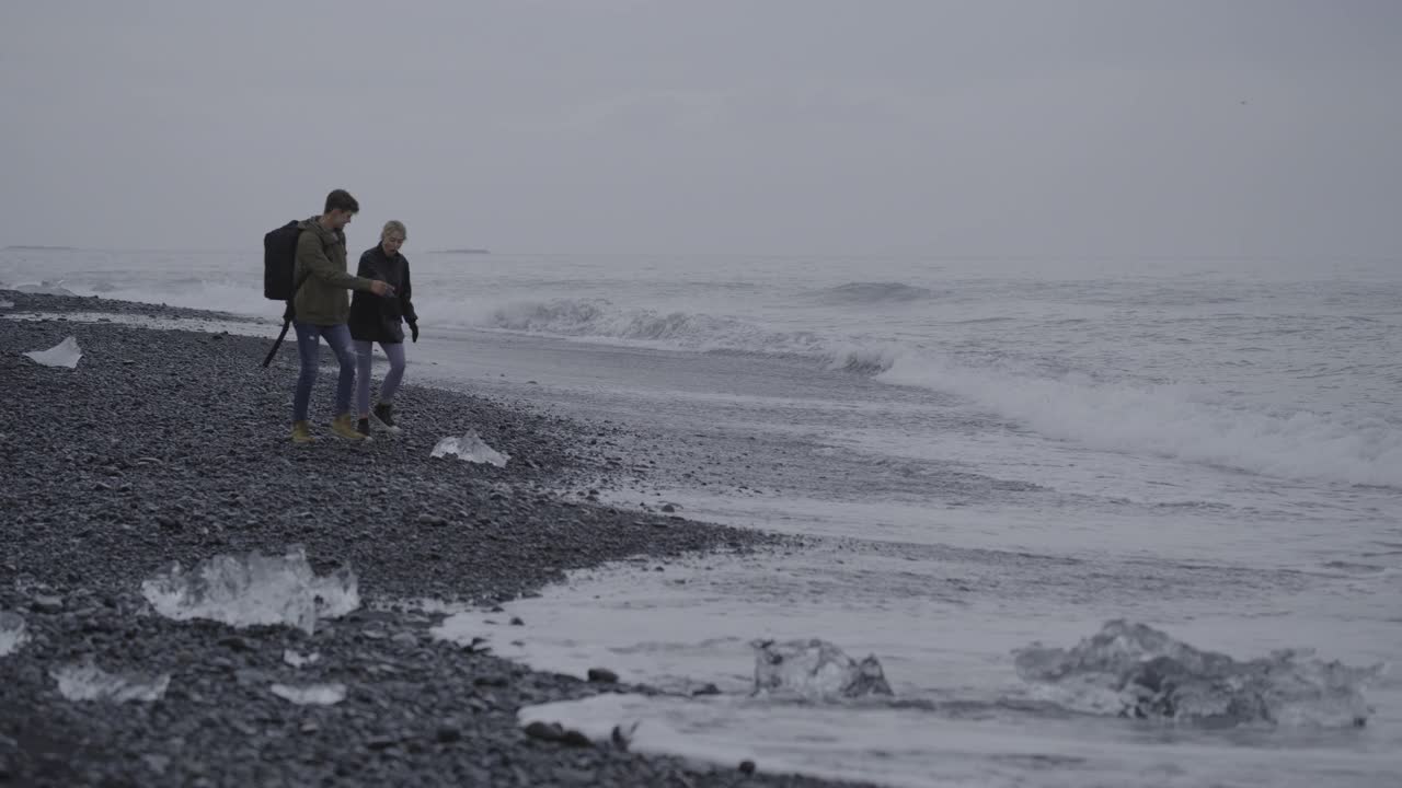 遥远的一对夫妇检查黑色沙滩上的大块冰/冰岛视频下载