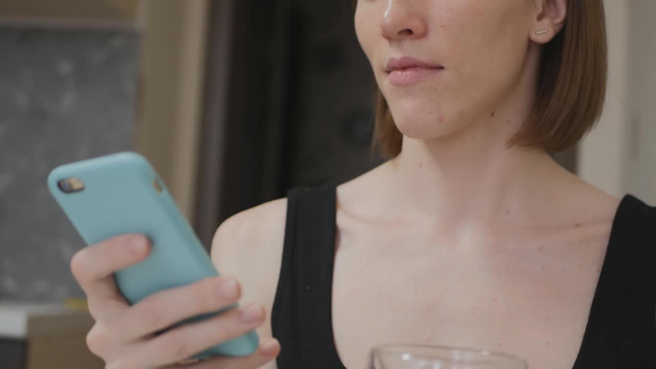 一个年轻漂亮的棕色头发的白种人女人正在用蓝绿色的手机输入信息，用双杯的杯子喝着饮料。早餐和好心情。视频素材
