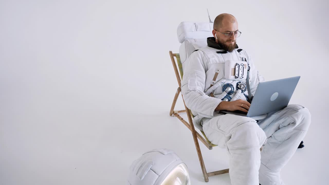 戴眼镜的宇航员坐在椅子上工作，膝盖上放着一台笔记本电脑。工作室视频下载