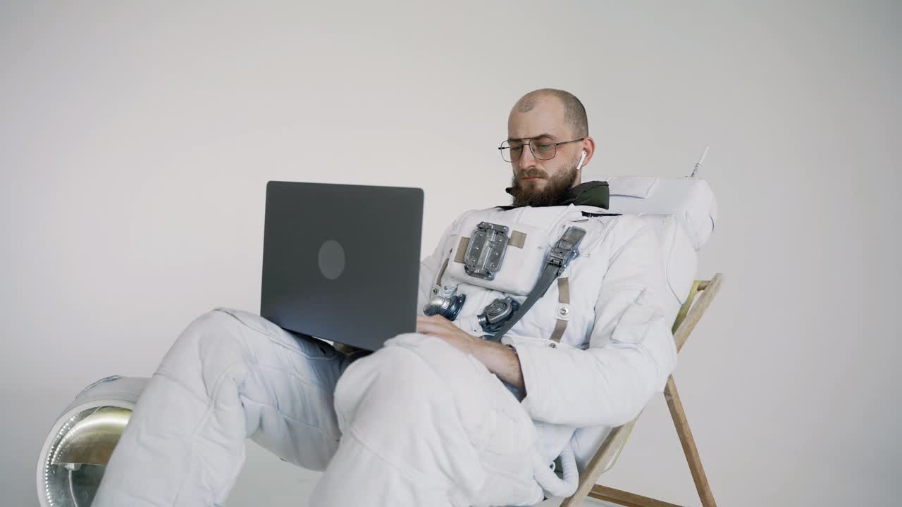 戴眼镜的宇航员坐在椅子上工作，膝盖上放着一台笔记本电脑。工作室视频素材
