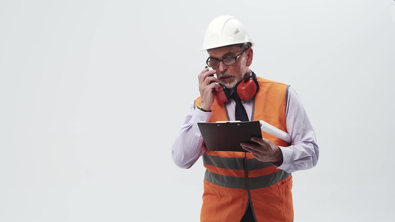 成年工程师穿着工作背心，戴着头盔，戴着眼镜和耳机，用对讲机说话，手里拿着平板电脑。穿着制服的人读着纸片，通过无线电传送信息视频素材