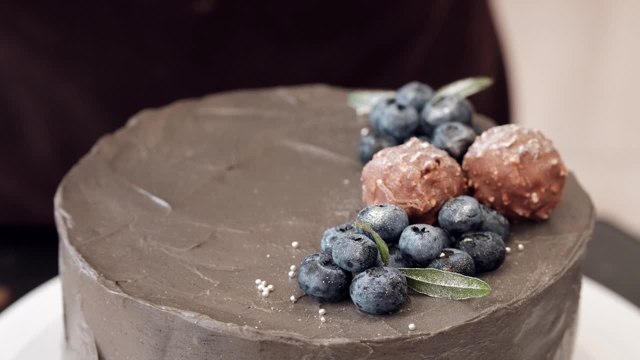 女人糖果师用浆果、巧克力和银珠子装饰灰色蛋糕，特写镜头。缓慢的运动。蛋糕制作过程。视频下载
