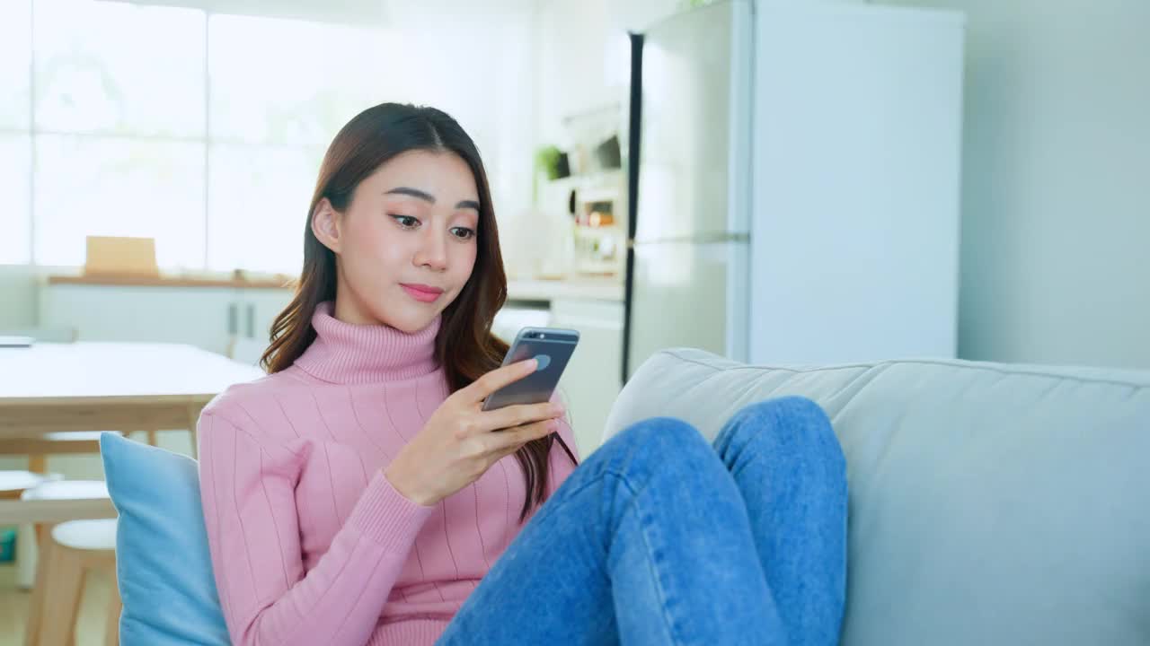 亚洲美女坐在沙发上，在家里讲手机。快乐迷人的年轻女孩在家休闲，放松身心，享受在客厅用智能手机交流讨论。视频素材