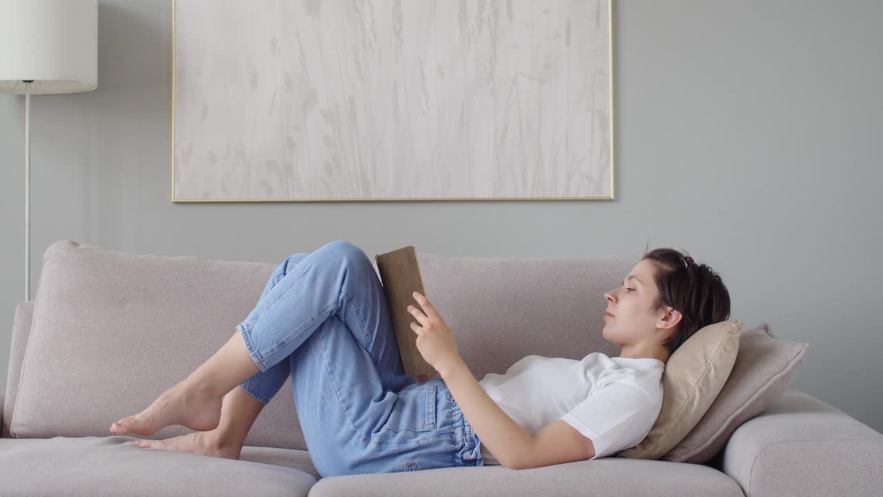 白人女孩阅读纸书放松在家里公寓自然光。漂亮女人躺在沙发上消磨闲暇时间拷贝空间文字。缓慢的运动。舒适、平静、闲暇，而冠状病毒视频素材