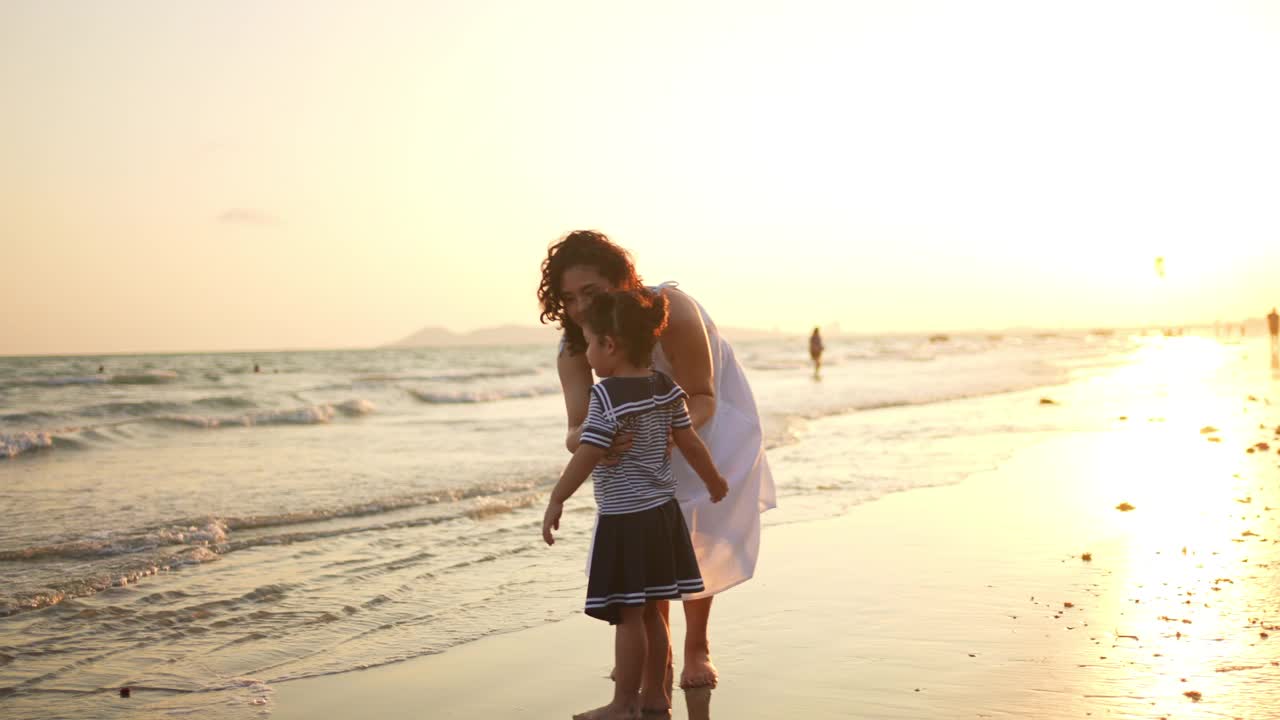 4K亚洲母亲玩和抱着可爱的女儿在沙滩上，在夏天的日落。视频素材