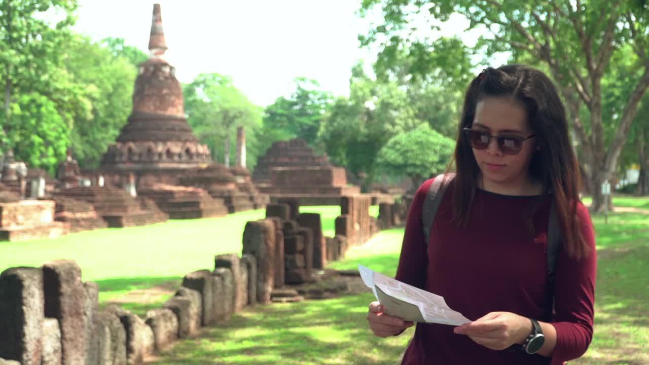 一位年轻的泰国女子提着一个小袋子，里面有一张纸地图，在树影下行走，探索著名的甘榜佛寺的历史时刻，前面的景色跟随，视频素材