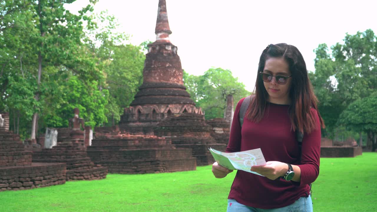 一名年轻的泰国女子带着一个小袋子，带着一张纸地图，在著名的贡彭Phet古庙探索历史宝塔时刻，视频素材
