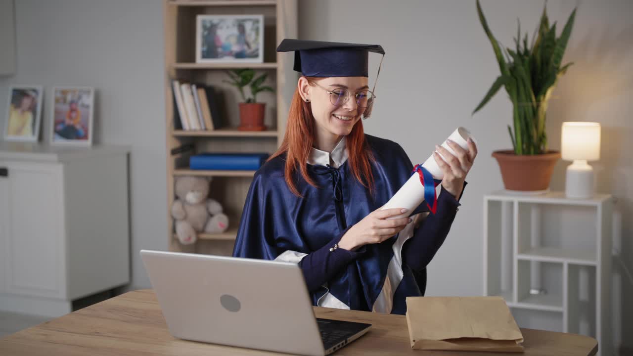 远程在线教育，年轻的女学生大学毕业生穿着学位服，手里拿着毕业证书，坐在家里用笔记本电脑背景的气球欢庆毕业和虚拟庄严的仪式视频下载