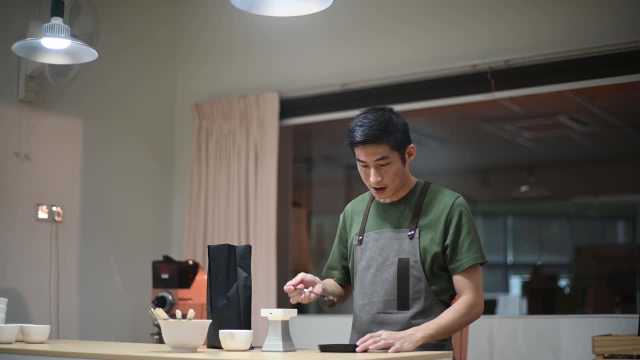 专业亚裔华裔男咖啡师，在明亮的房间里准备咖啡杯，取样品，舀咖啡，用秤称重视频素材