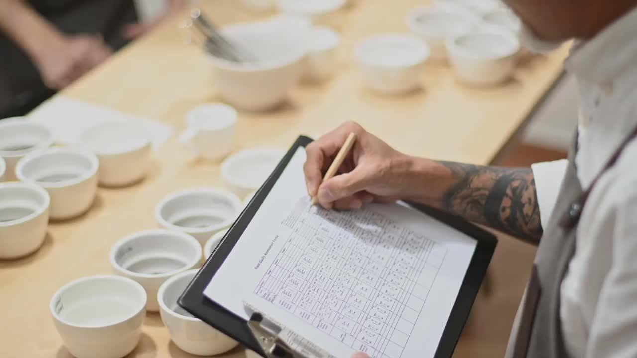 高角度的视角，专业的亚洲华人咖啡师评分者表演咖啡杯，品尝咖啡杯后在剪贴板上写下视频素材
