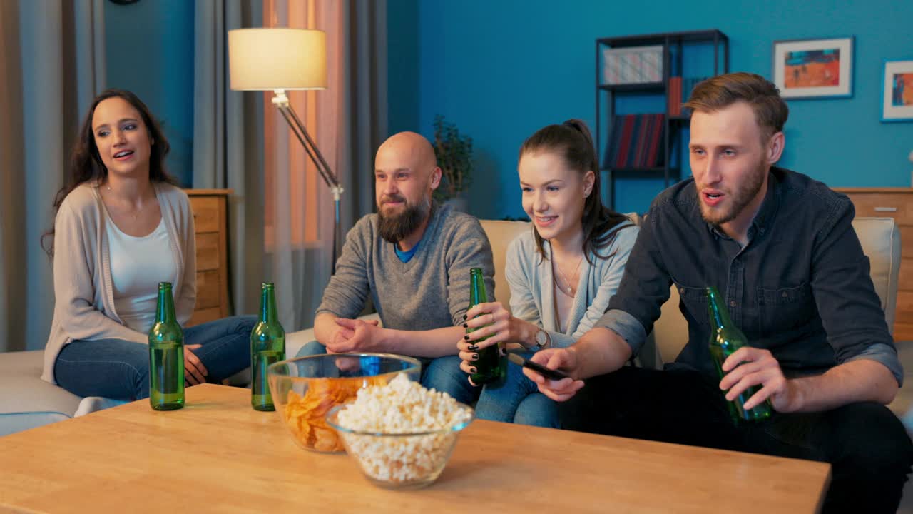四个好朋友在彼此的陪伴下度过了一个晚上，坐在客厅的沙发上，喝着啤酒，男人拿着电视遥控器打开了歌舞表演，大家都饶有兴趣地看着，笑着视频下载