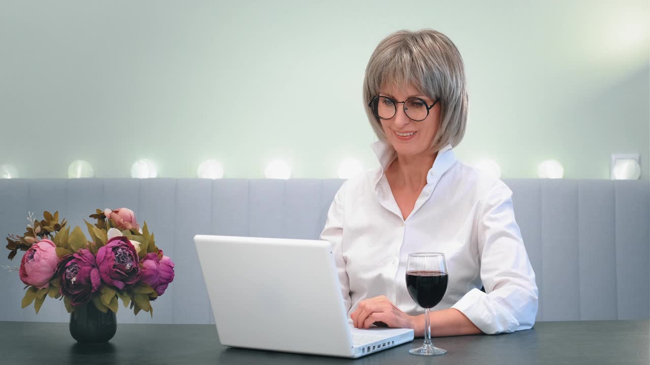 一位快乐美丽的老妇人喝着酒打电话给朋友，在笔记本电脑上看亲戚的视频。一位满头白发的老妇人面带微笑，手里拿着杯子敬酒，在网上聊天视频素材