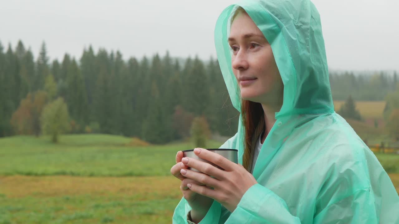 秋雨中，穿着雨衣的轻松女子在大自然中喝茶视频下载