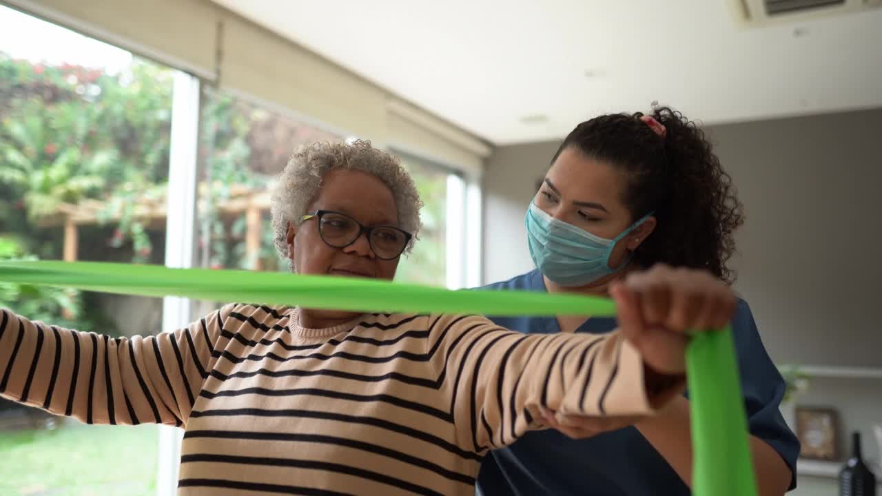 老年妇女在物理治疗师的支持下，戴着防护口罩，用阻力带做运动视频下载