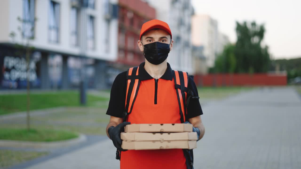 照片中的快递员背着红色背包，手里拿着装在纸箱里的披萨，戴着防护口罩和手套。优秀的交付。递送服务视频素材