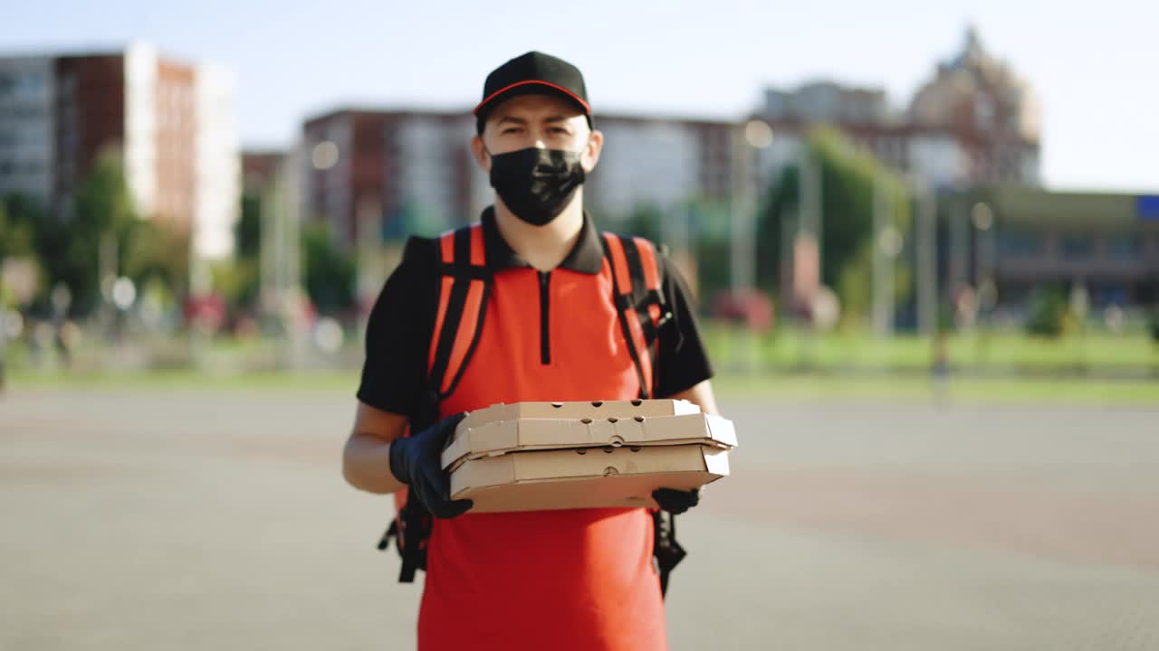照片中的快递员背着红色背包，手里拿着装在纸箱里的披萨，戴着防护口罩和手套。交付服务。优秀的交付视频素材