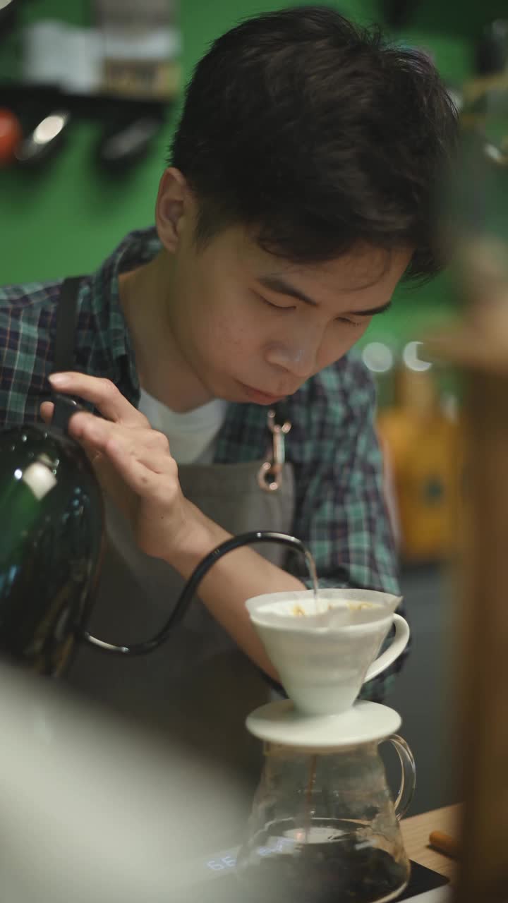 亚洲华人男咖啡师拿着水壶将热水倒在咖啡过滤器上，在咖啡店里准备滴咖啡视频素材