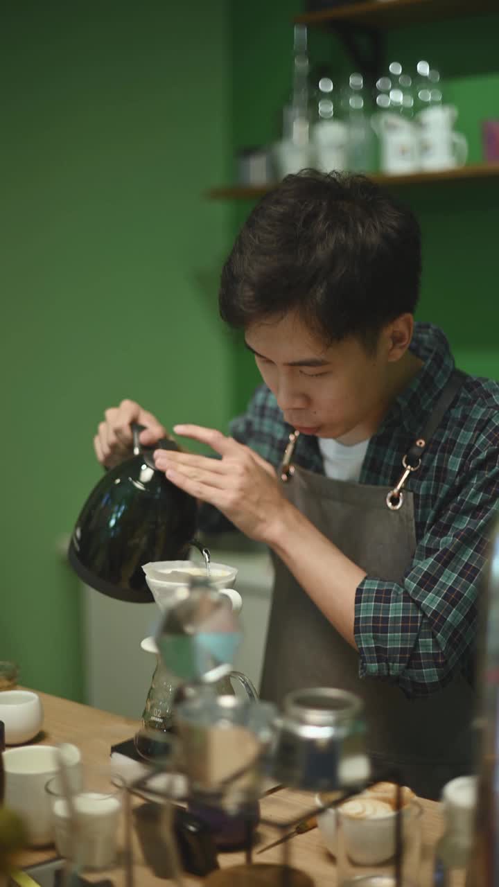 亚洲华人男咖啡师拿着水壶将热水倒在咖啡过滤器上，在咖啡店里准备滴咖啡视频素材