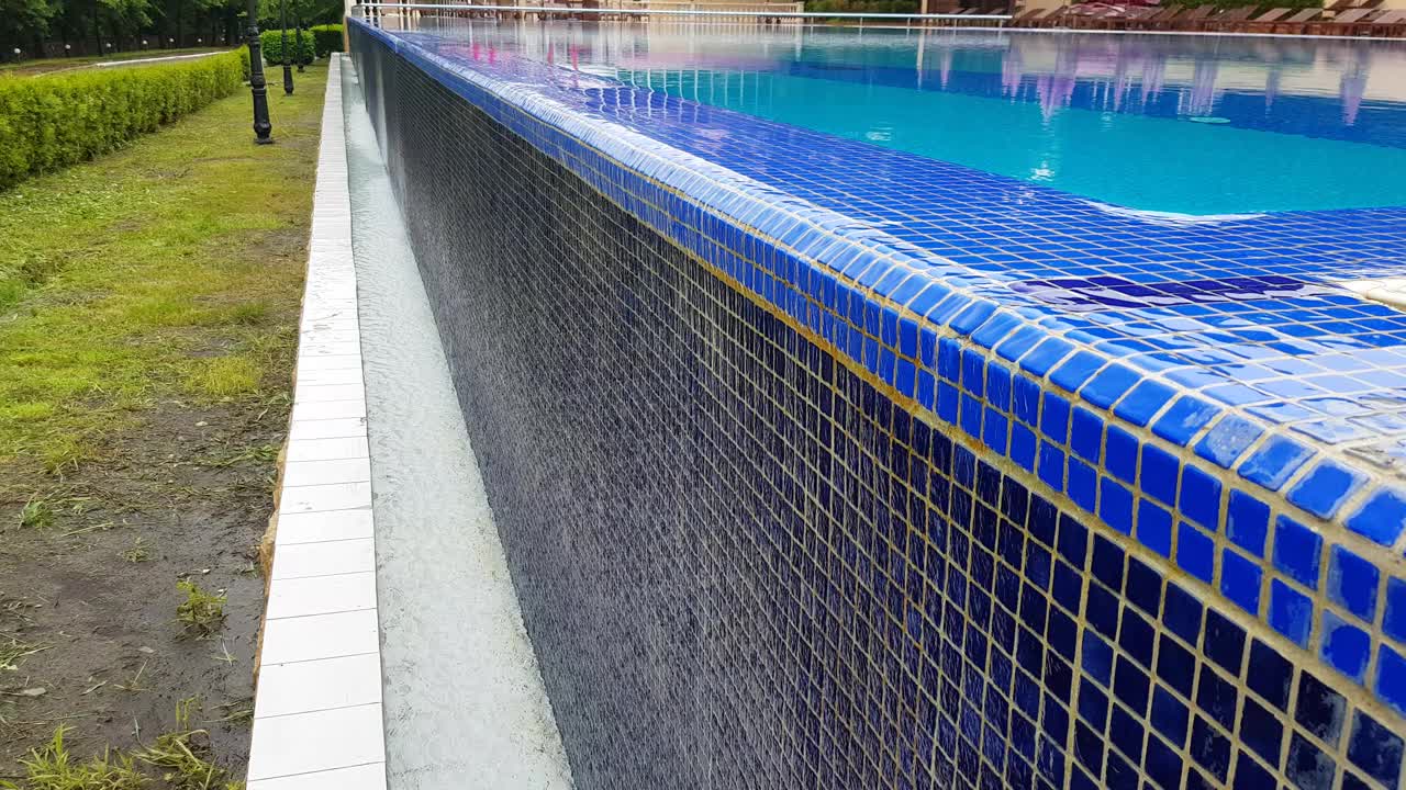 4k视频，一个空的室外游泳池的全景，蓝色的水面和蓝色的背景纹理的小马赛克瓷砖，水从室外游泳池流下墙视频下载