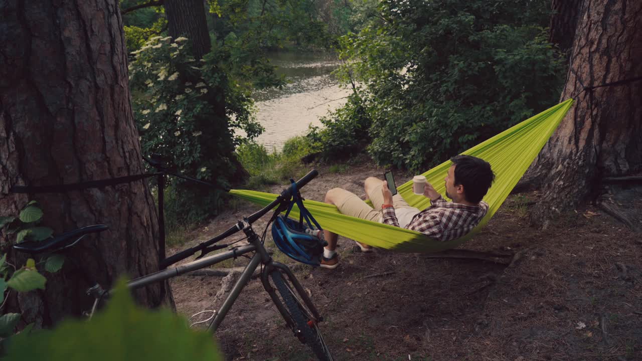 一名男子骑着自行车来到公园，在俯瞰湖面的吊床上休息，一边喝着咖啡一边用智能手机上网。人们旅行度假的概念视频下载