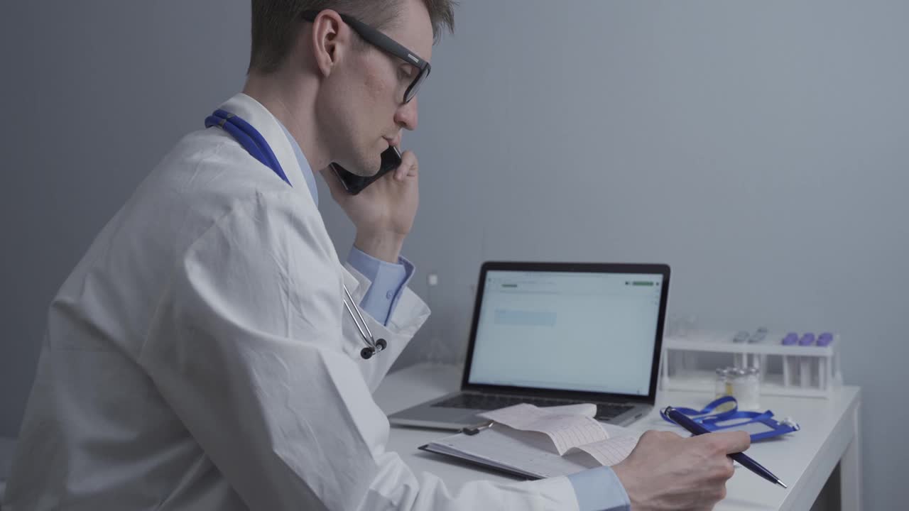 心脏科医生在诊所电脑旁的桌子旁检查病人的心电图，并通过电话咨询。医疗和药品。疾病的诊断和治疗视频下载