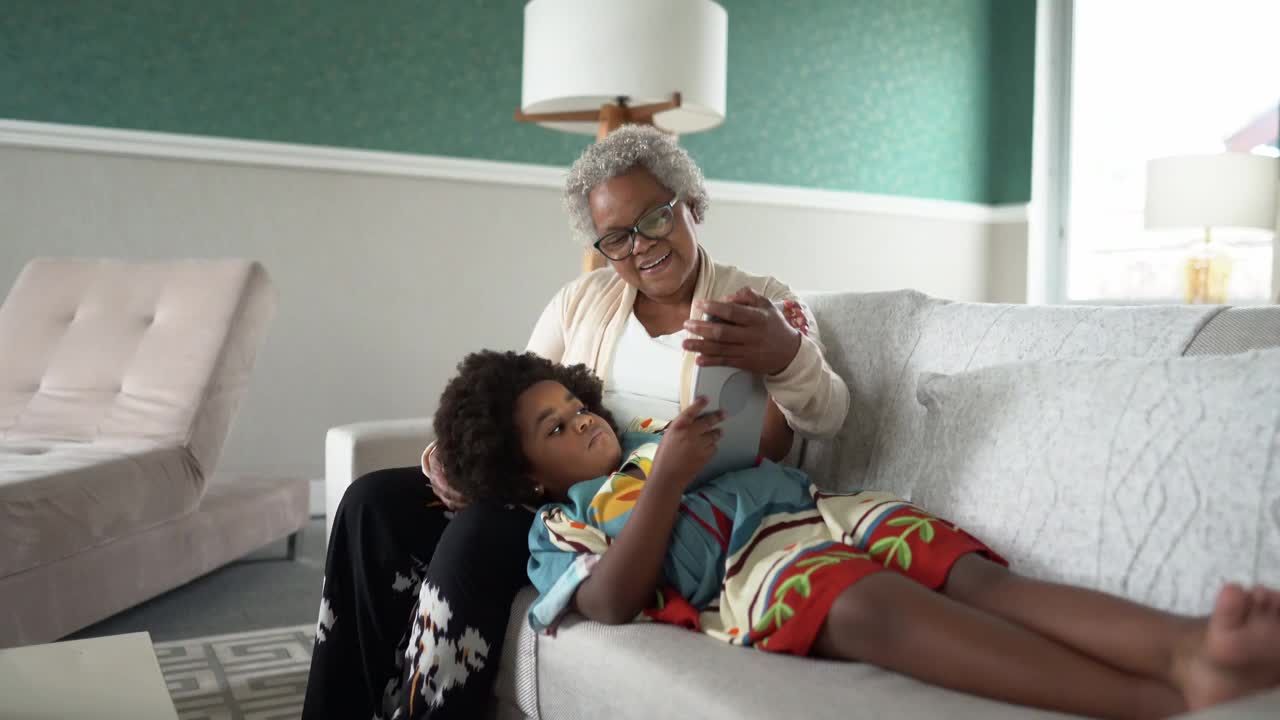 女孩躺在奶奶的腿上，在家里使用数码平板电脑视频下载