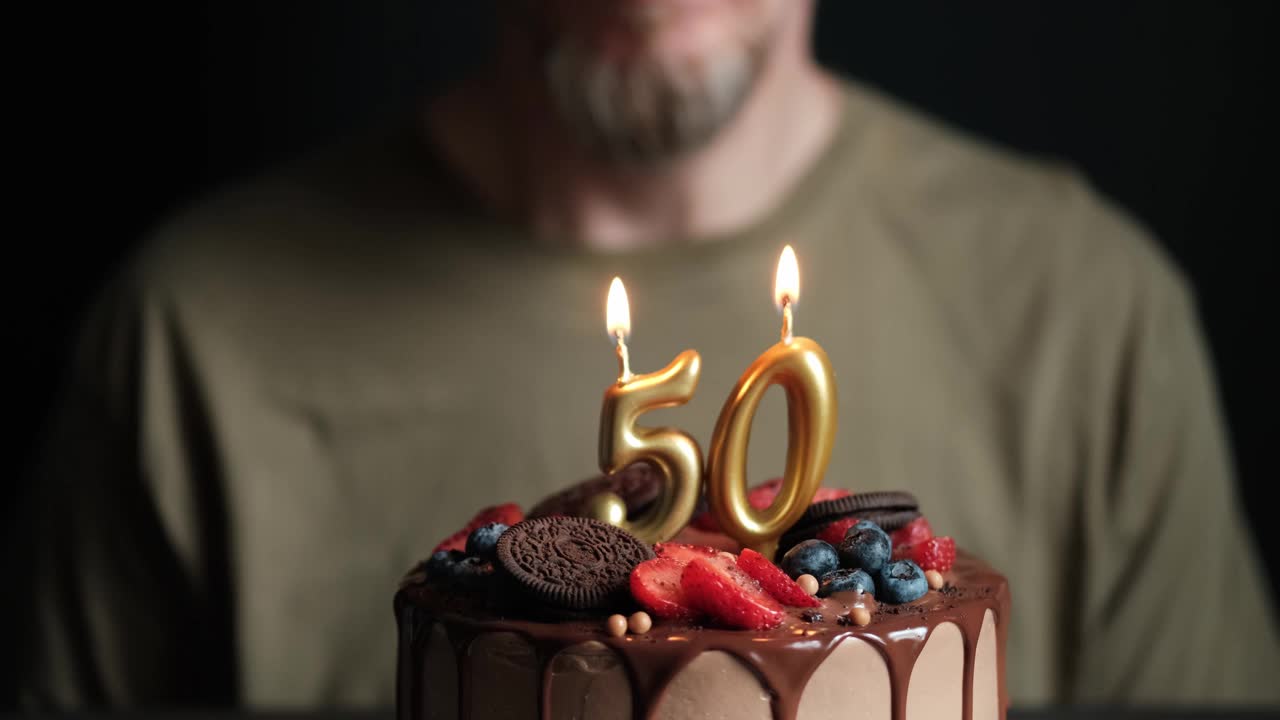 50岁的男人吹灭了巧克力蛋糕上燃烧的50支蜡烛。50周年生日蛋糕。慢动作视频下载