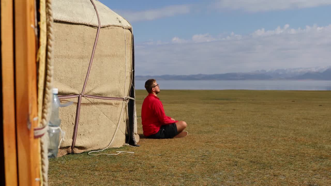 在吉尔吉斯斯坦，一个穿着红色衣服的旅行者坐在可以看到宋库尔湖和山脉的蒙古包营地，迎接明媚的早晨视频素材