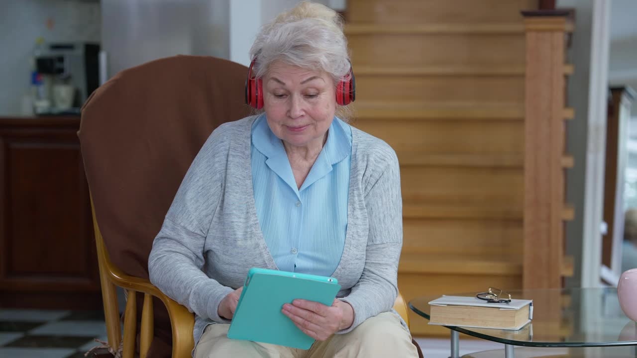 家中客厅舒适的扶手椅上，戴着耳机享受音乐的快乐悠闲的女退休人员肖像。积极的白种老妇人微笑着在室内跳舞视频素材