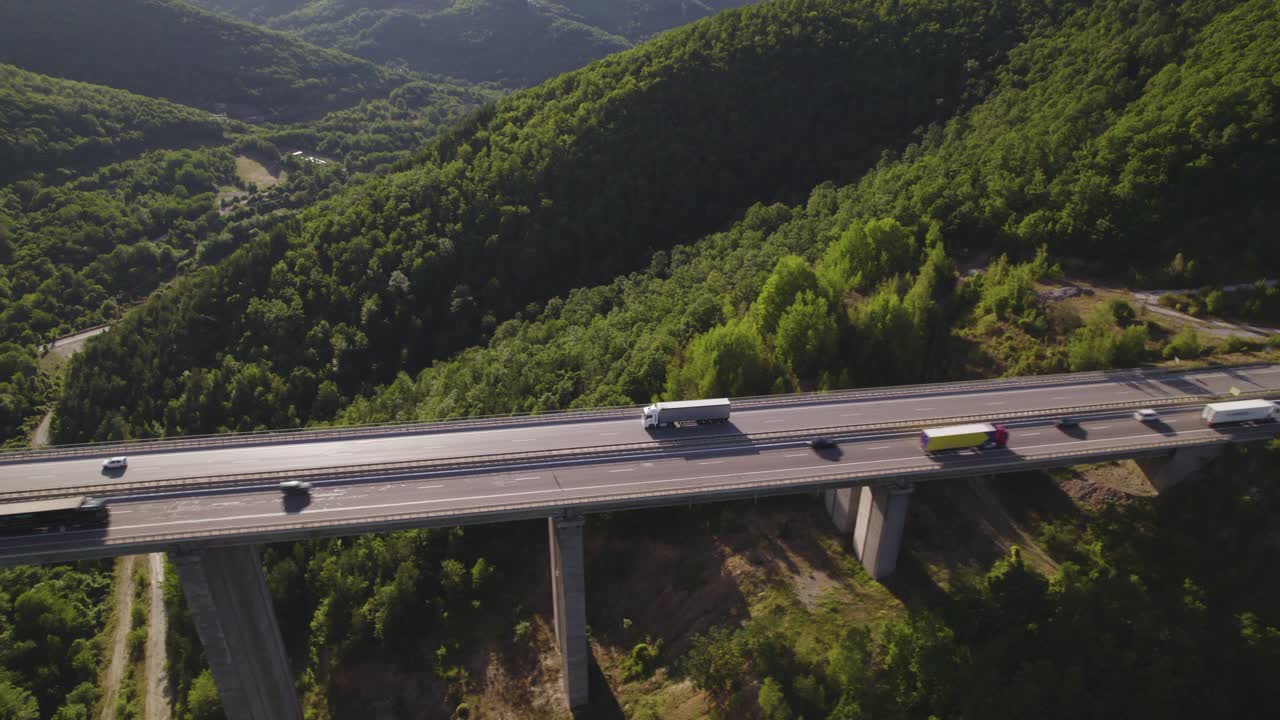 鸟瞰图白色货运半挂车行驶在两路高速公路高架桥上的绿色山丘视频素材