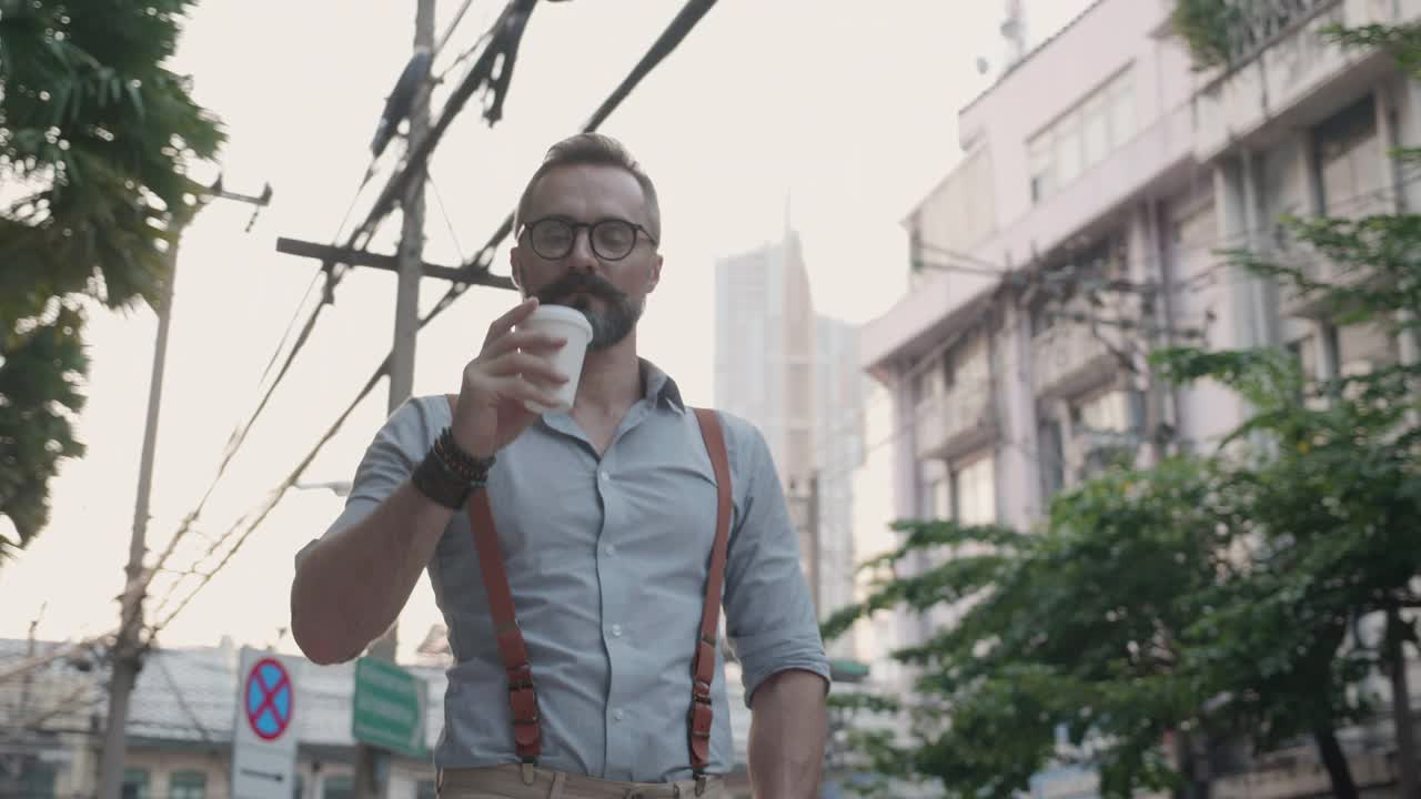 英俊男子的肖像站在街上自信地喝着咖啡或饮料，胡须男子自信地拿着一杯饮料放松在城市，户外。视频素材