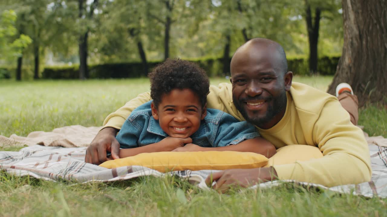 快乐的黑人男子和他的小儿子躺在公园的草坪上视频素材